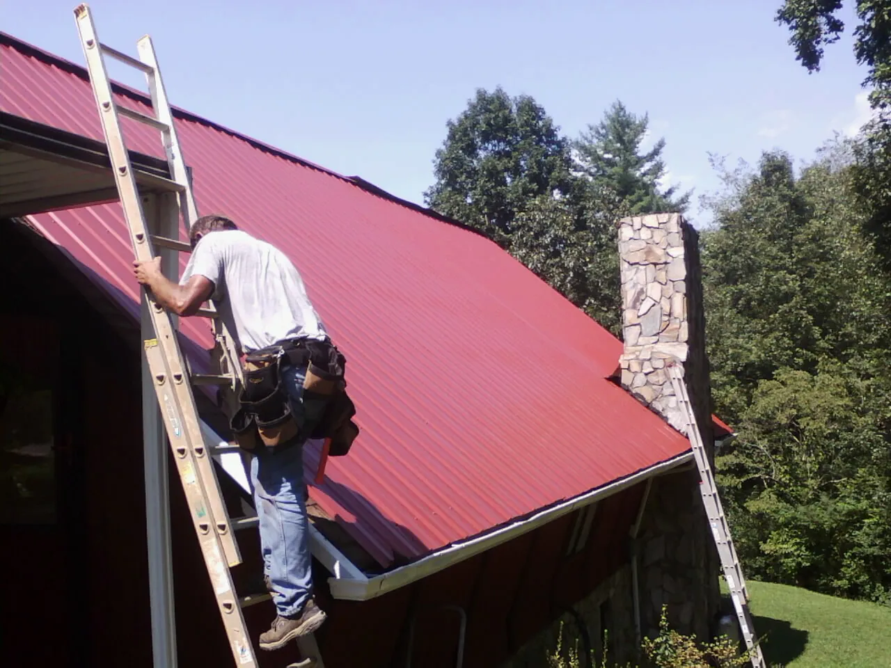 Roofer on a ladder beside a red metal roof and stone chimney — Soffit & Fascia Repair by Roofing Brothers in Creston